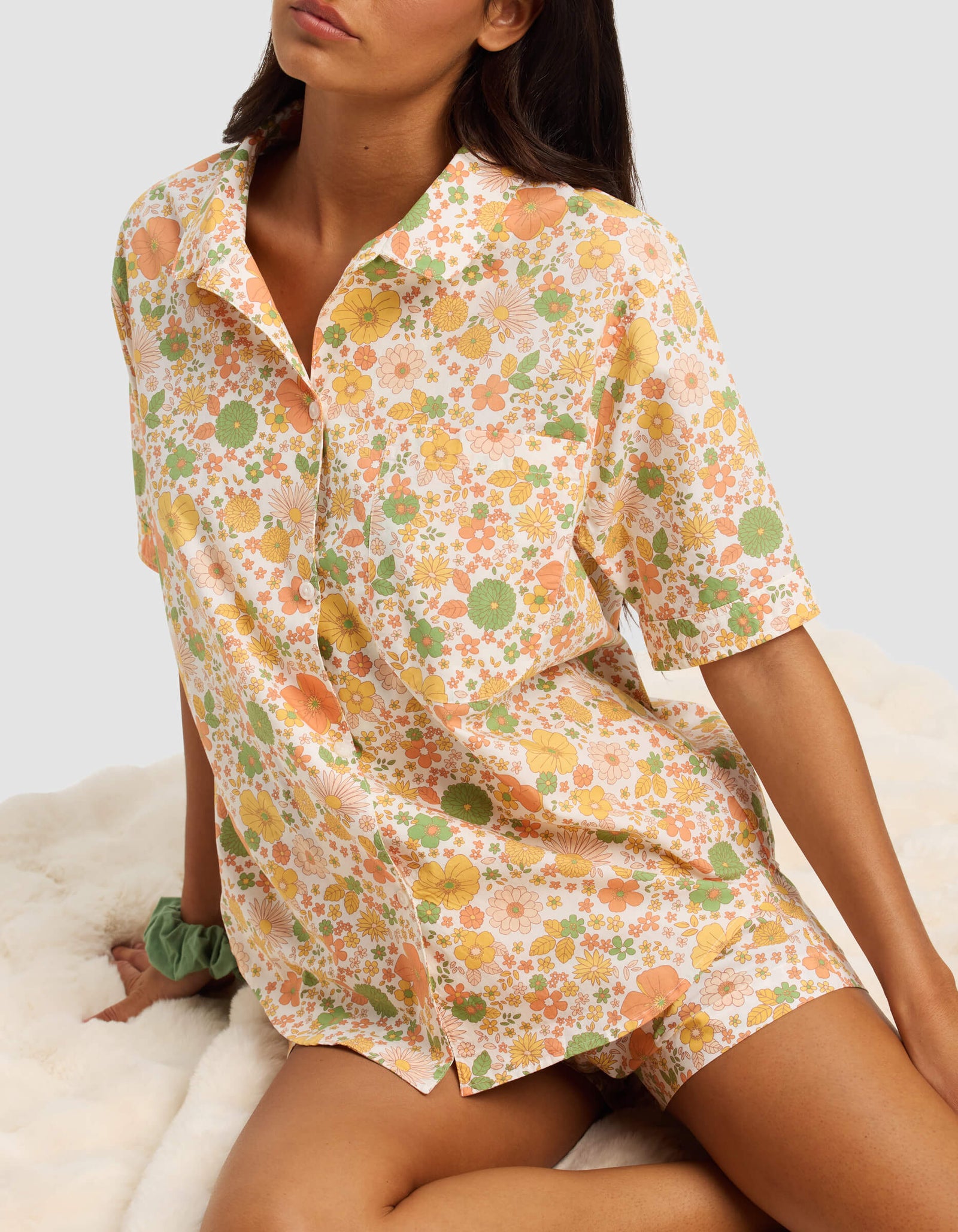 A woman sits on a fluffy white surface wearing the Cozy Earth Women's Sutton Short Sleeve Pajama Top with a pastel floral pattern in orange, green, and yellow, and has a green scrunchie on her wrist.