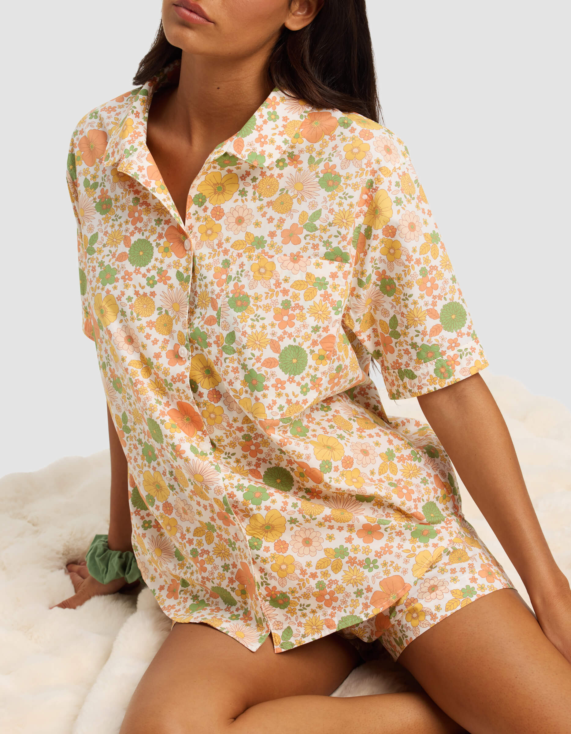A woman sits on a fluffy white surface wearing the Cozy Earth Women's Sutton Short Sleeve Pajama Top with a pastel floral pattern in orange, green, and yellow, and has a green scrunchie on her wrist.|Color:Wildflower