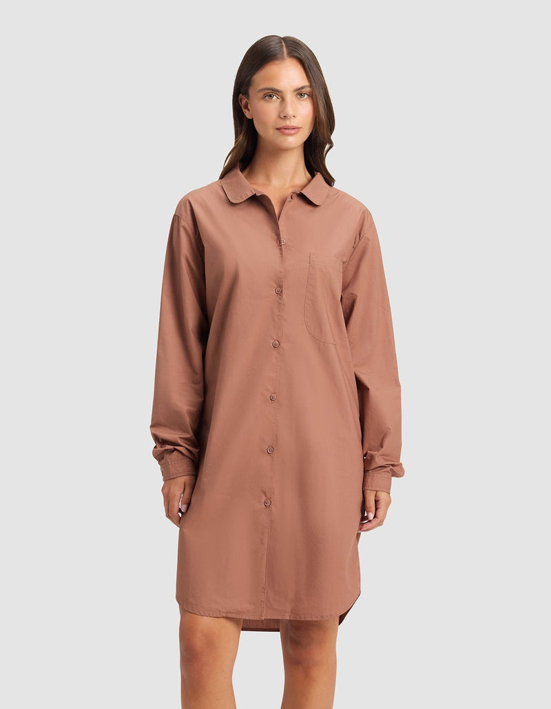 A woman stands against a plain background wearing the Cozy Earth Women's Sutton Sleep Dress, a loose, long-sleeve, button-down brown shirt dress with a collar and chest pocket.