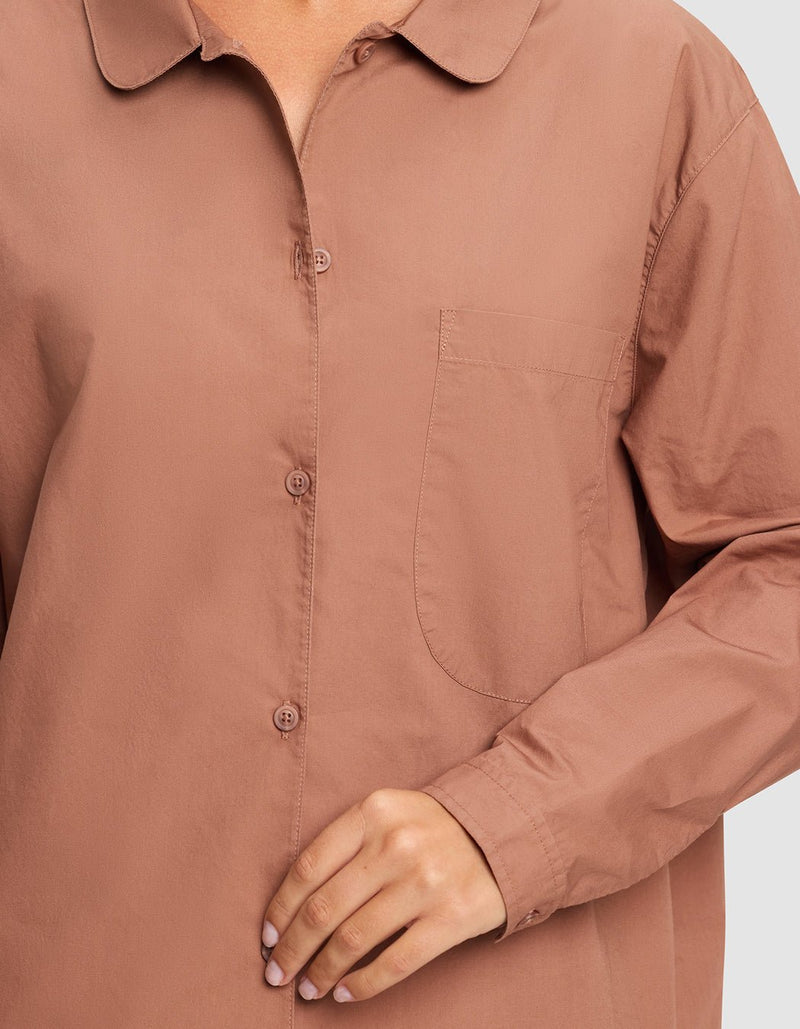 A person shown from shoulders to hips wears the Cozy Earth Women's Sutton Sleep Dress, a loose, long-sleeve, button-up sleep shirt in muted brown with a front pocket. Their hand rests near the buttons.