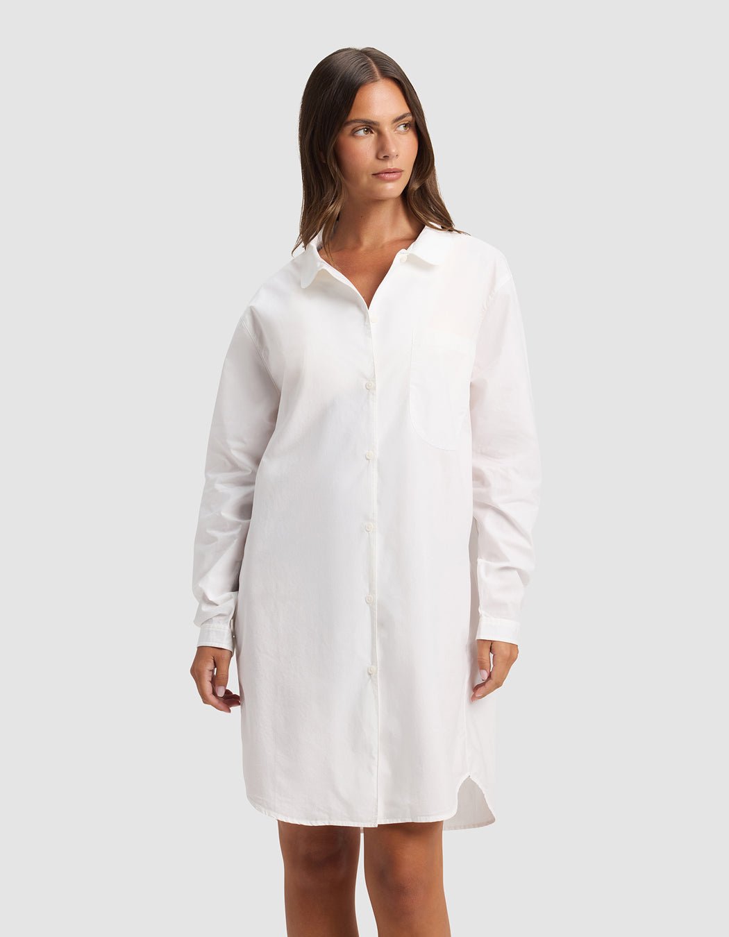 A woman wearing the Cozy Earth Women's Sutton Sleep Dress, a long-sleeved, button-down white shirt dress, stands against a plain light gray background and looks slightly to the side.|Color:White