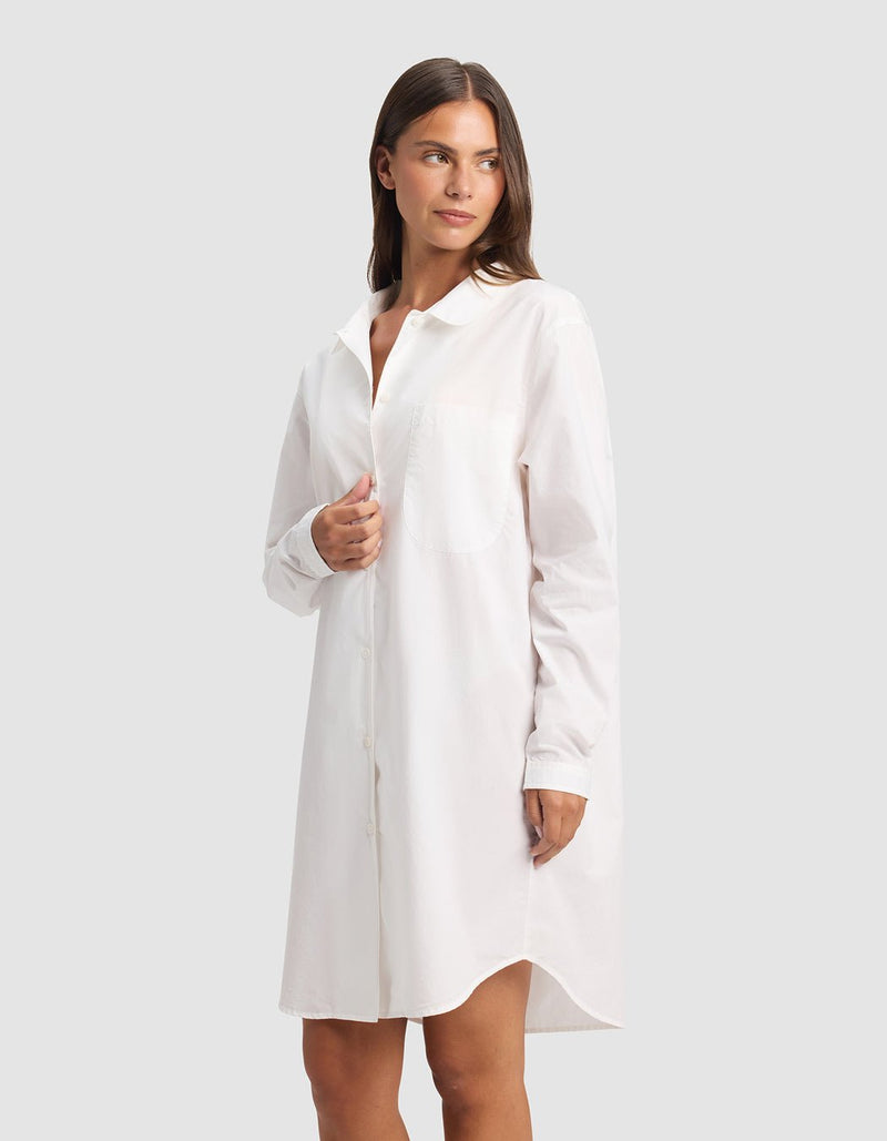 A woman in the Cozy Earth Women's Sutton Sleep Dress, a long-sleeved white button-down style, stands against a plain light gray background, gazing slightly left with a relaxed expression.