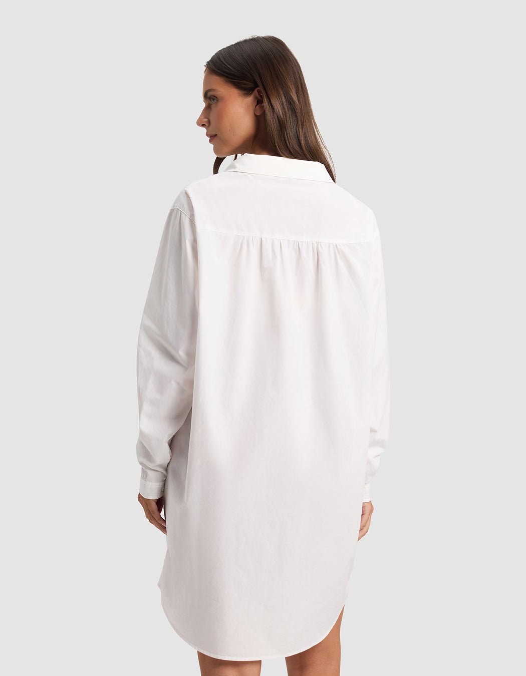 A woman with long brown hair faces away, wearing the Cozy Earth Women's Sutton Sleep Dress—a loose-fitting white, long-sleeve shirt dress—set against a plain light gray background.