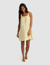 A woman with long, dark hair models the Cozy Earth Women's Sutton Tank Sleep Dress—a loose, sleeveless, light yellow nightdress with a gathered neckline and bow detail—standing barefoot against a plain light background.|Color:Meringue