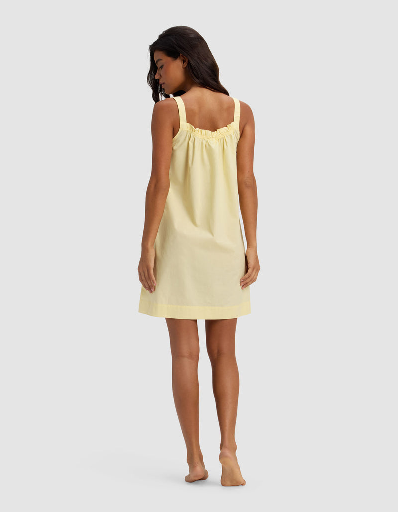 A woman with long dark hair stands barefoot, facing away, wearing the Cozy Earth Women's Sutton Tank Sleep Dress in light yellow, featuring a sleeveless, loose fit and gathered upper back detail, against a plain light gray background.