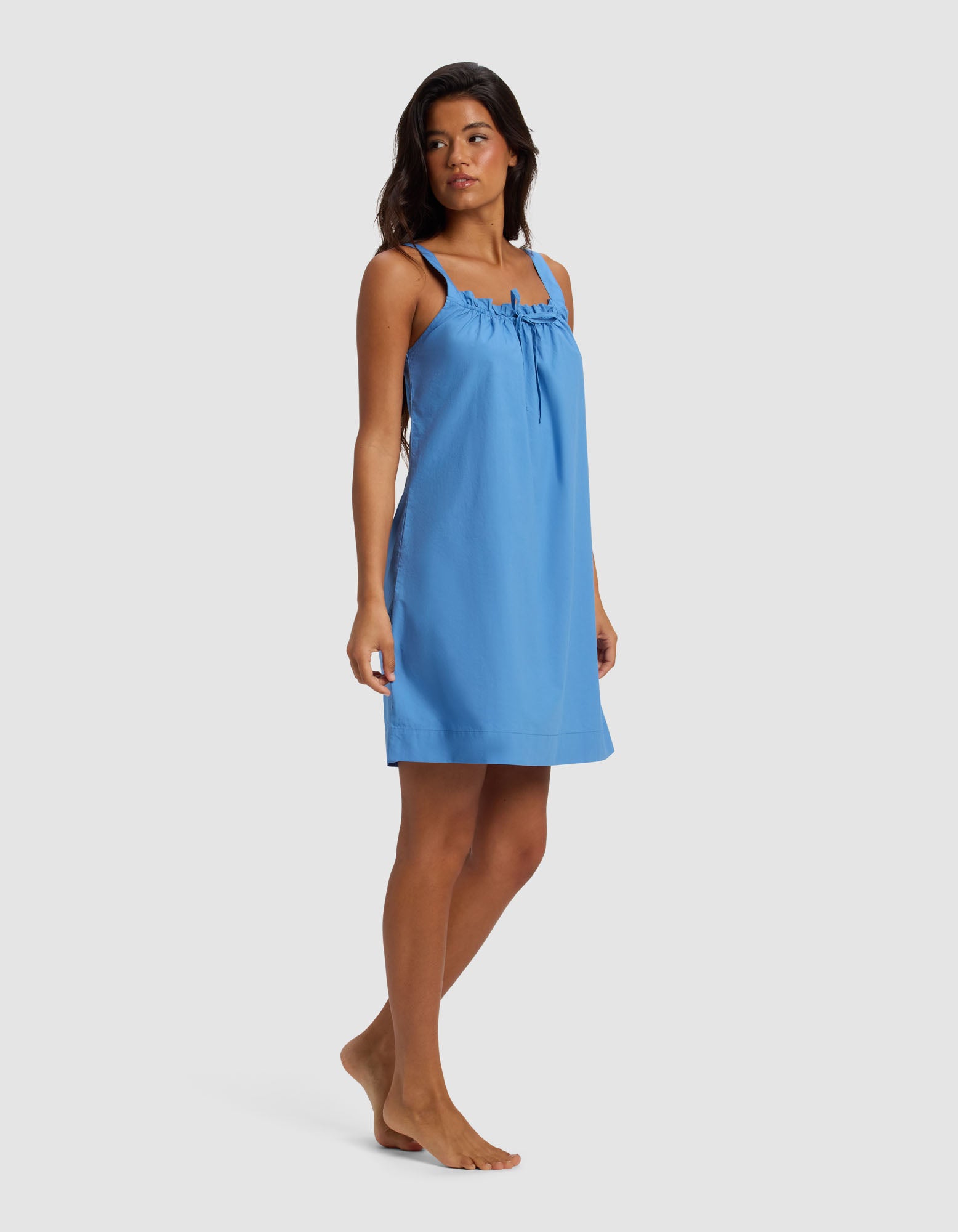 A woman stands barefoot on a plain background, wearing the Cozy Earth Women's Sutton Tank Sleep Dress—a sleeveless, light blue knee-length dress with thin straps and a gathered neckline—while looking slightly to the side.