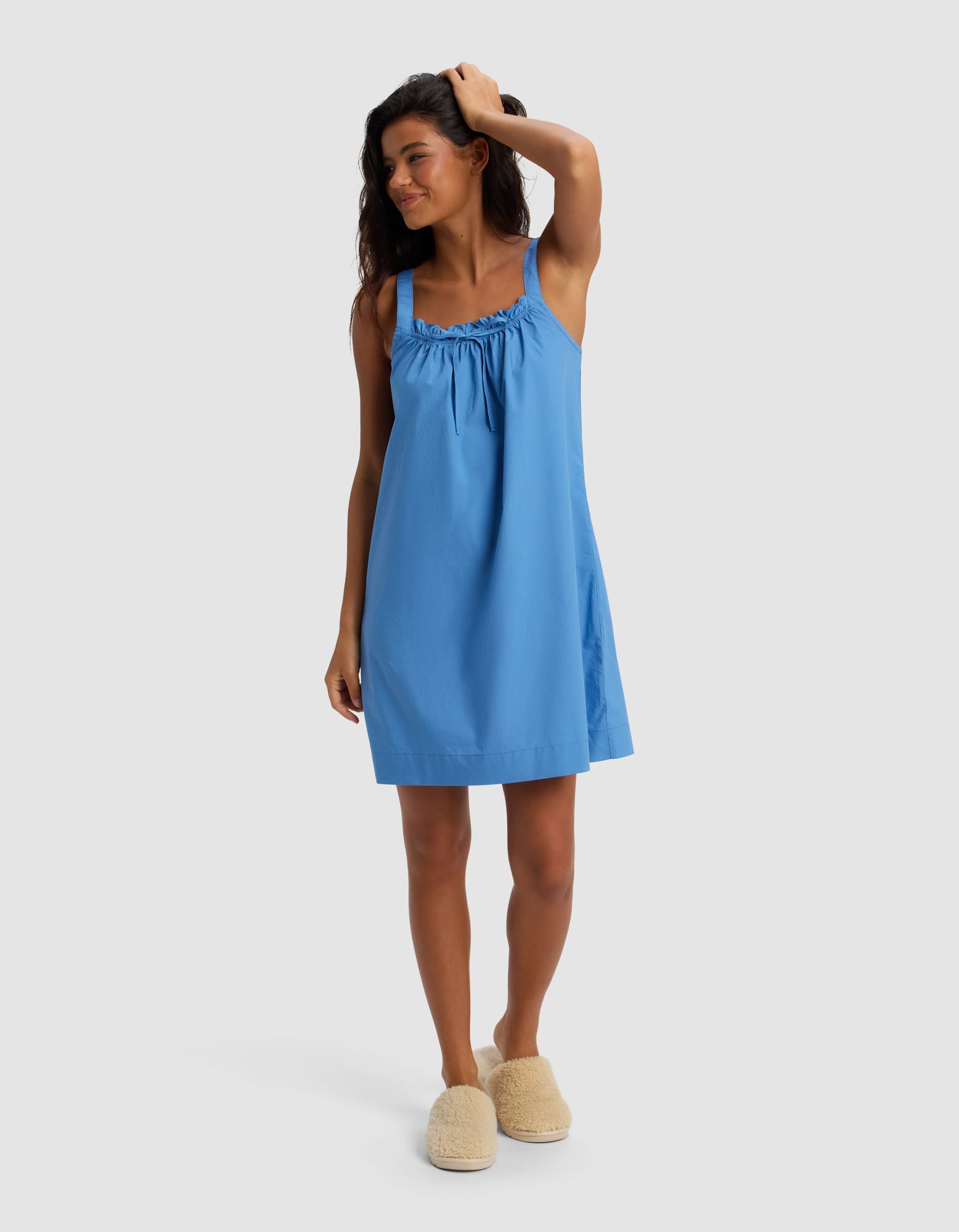 A woman wearing Cozy Earth's Women's Sutton Tank Sleep Dress in blue stands barefoot on a white background, smiling with one hand in her hair and wearing fuzzy beige slippers.|Color:Sailor