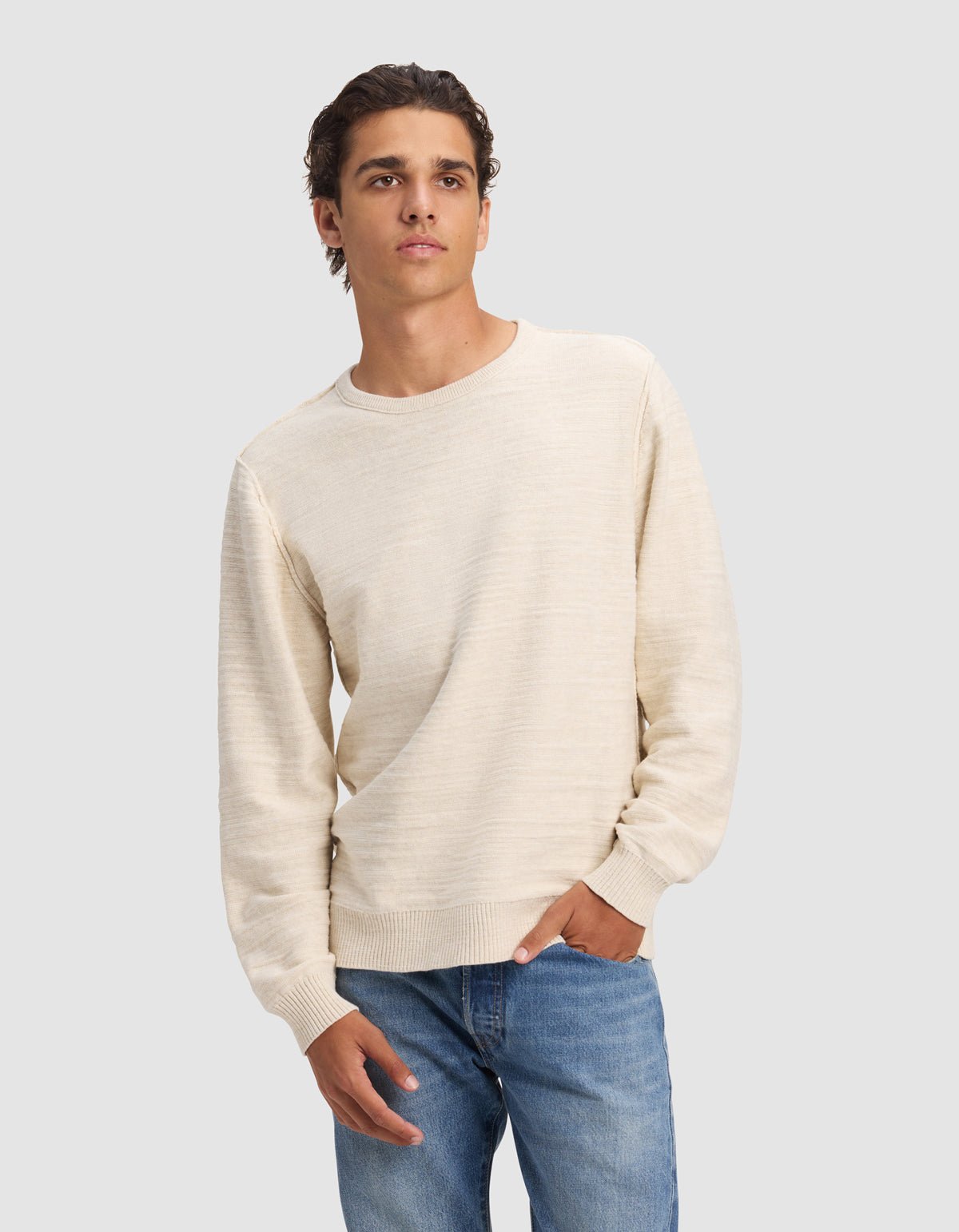 A young man models the Cozy Earth Men's Tate Crewneck in light beige with blue jeans, standing against a plain gray background and looking to the side, one hand in his pocket.