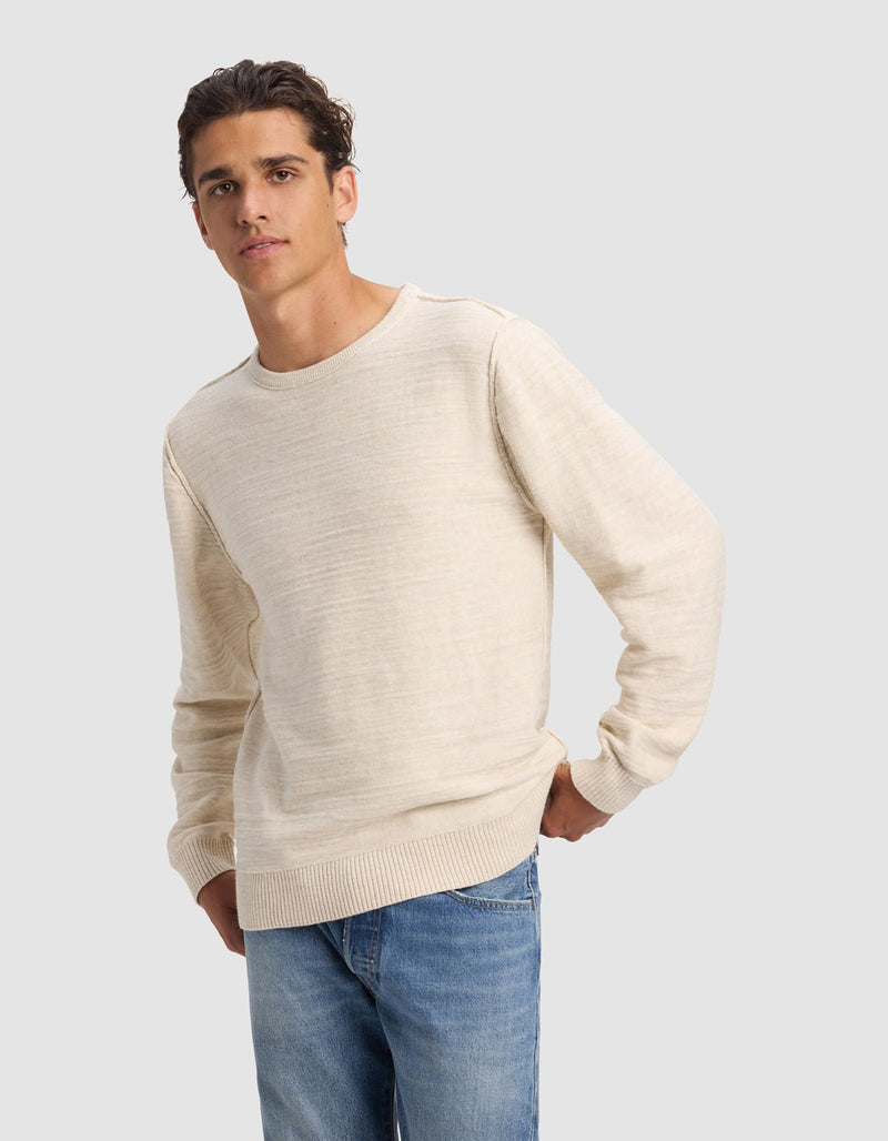 A young man wears the Cozy Earth Men's Tate Crewneck in light beige with blue jeans, standing against a plain gray background with one hand on his hip and a neutral expression.