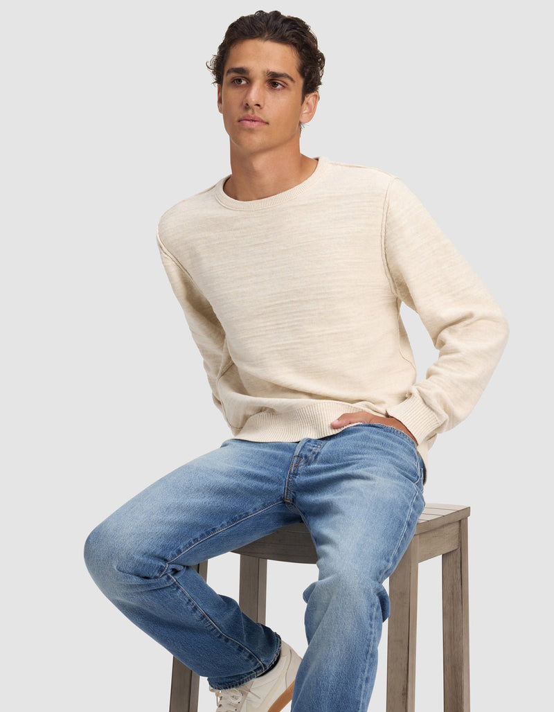 A young man with short curly hair wears the Cozy Earth Men's Tate Crewneck and blue jeans while seated on a wooden stool against a plain light gray background, gazing slightly to the side.