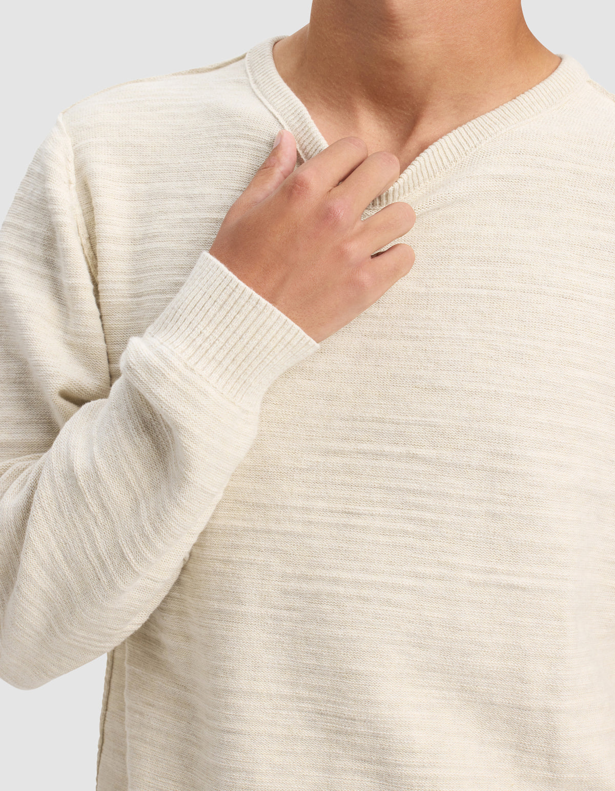 A man models the Cozy Earth Men's Tate Crewneck, a cream-colored knit sweater with long sleeves, shown from shoulders to waist against a plain light gray background. One hand touches the collar near his neck.