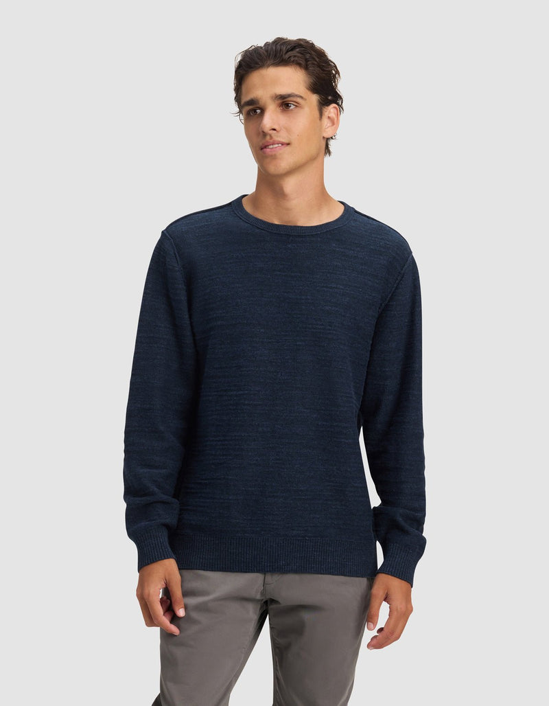 A young man with short, wavy brown hair wears the Cozy Earth Men's Tate Crewneck in dark blue and light gray pants, standing against a plain light gray background with a neutral expression, gazing slightly to the side.