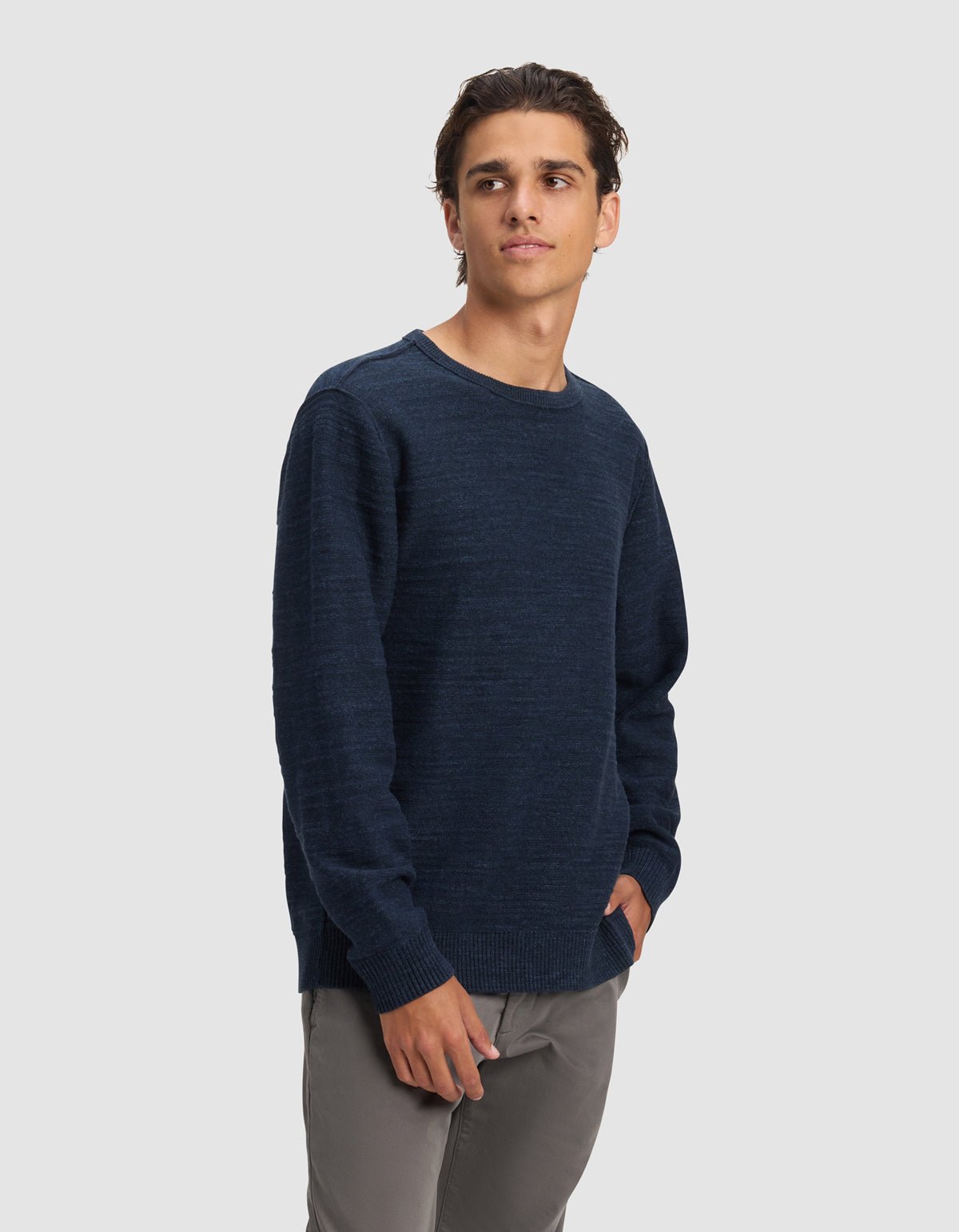 A young man with dark hair wears the Cozy Earth Men's Tate Crewneck in navy blue and gray pants, standing against a plain light gray background, gazing slightly left with his right hand in his pocket.|Color:Eclipse