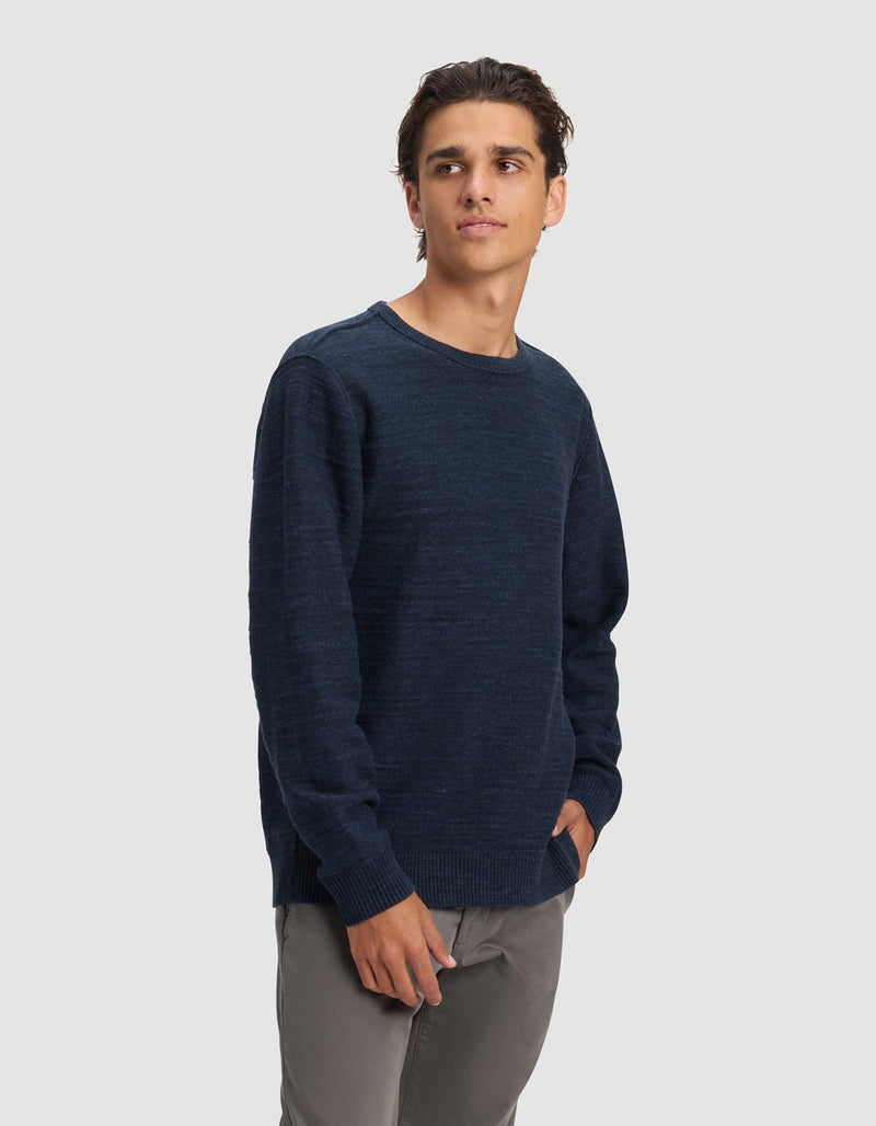 A young man with dark hair wears the Cozy Earth Men's Tate Crewneck in navy blue and gray pants, standing against a plain light gray background, gazing slightly left with his right hand in his pocket.