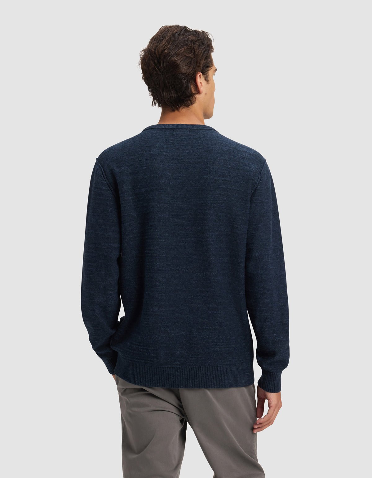 A man stands facing away from the camera, wearing Cozy Earth's Men's Tate Crewneck in dark blue and gray pants against a plain light gray background.