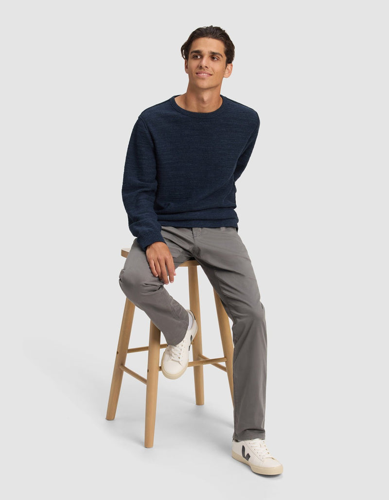 A young man with short, dark hair sits on a wooden stool against a plain background, relaxed and wearing the Cozy Earth Men's Tate Crewneck in navy blue, light gray pants, and white sneakers.