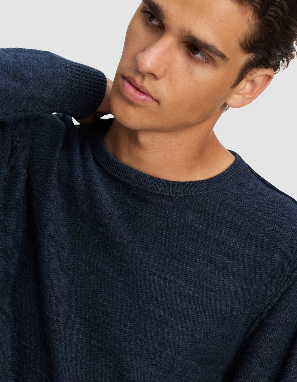 A young man with short brown hair, wearing the Cozy Earth Men's Tate Crewneck, looks off to the side with his hand behind his neck against a plain light background.