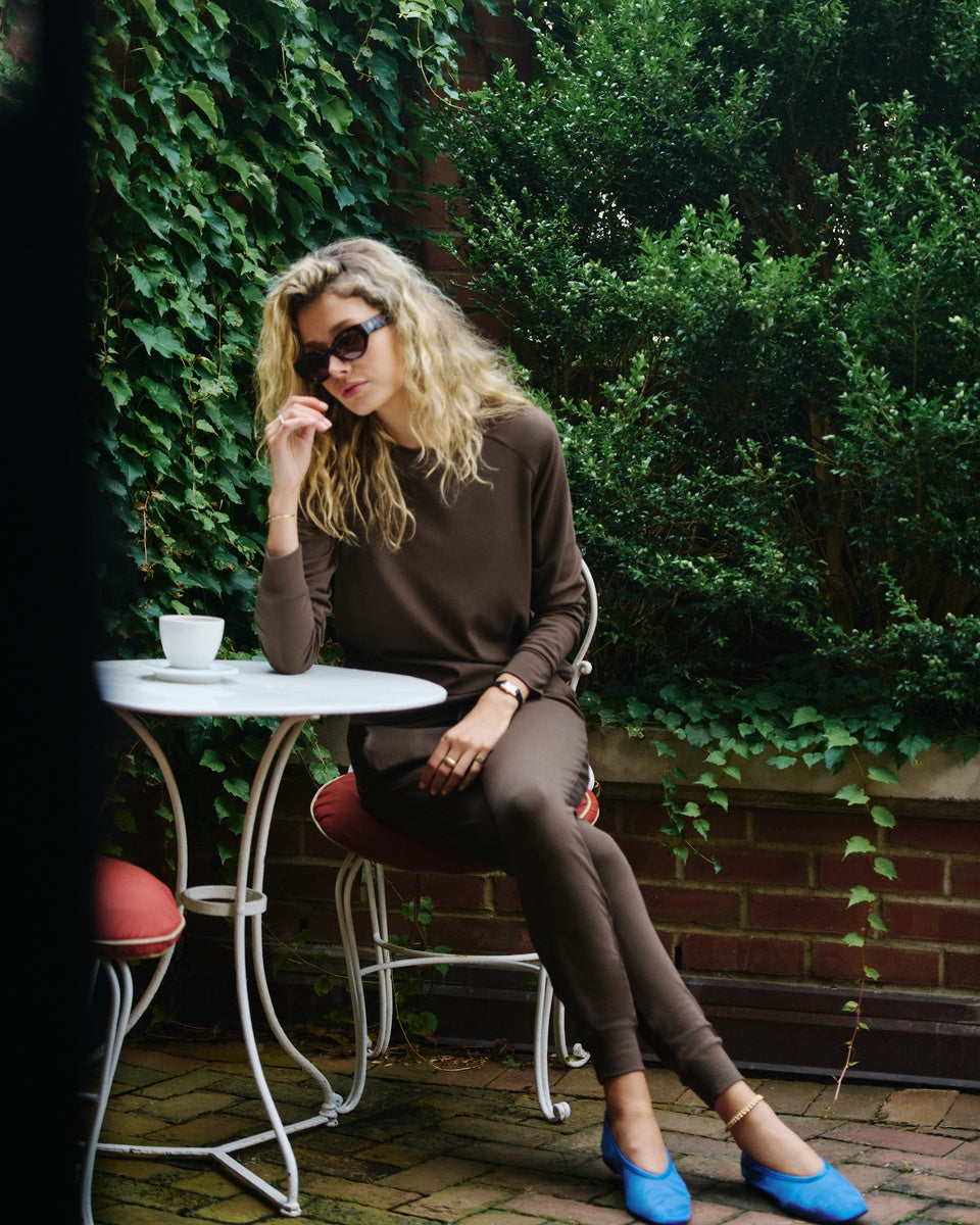 A woman with blonde hair and sunglasses sits at an outdoor patio table wearing a brown long-sleeve top and matching brown pants, with bright blue shoes, against a background of green ivy and brick. |Color:Espresso
