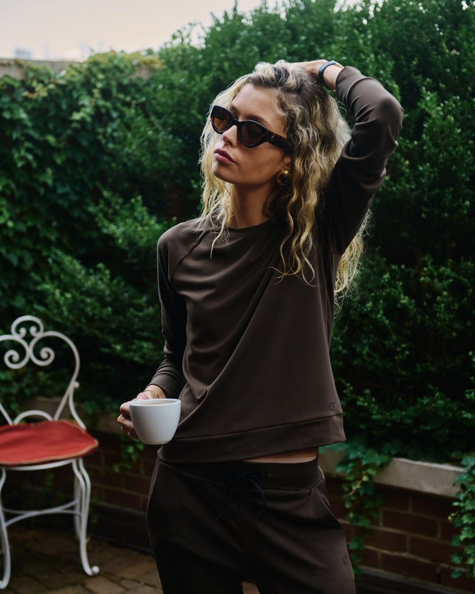 A woman wearing a brown Cozy Earth Brushed Bamboo Pullover and Jogger set, styled with black sunglasses and holding a white mug, standing outdoors in front of a green hedge. 
