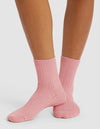 Close-up of a person wearing Cozy Earth's The Plush Modern Crew Sock in light pink, standing on a white surface with legs crossed at the ankles.|Color:Tulip