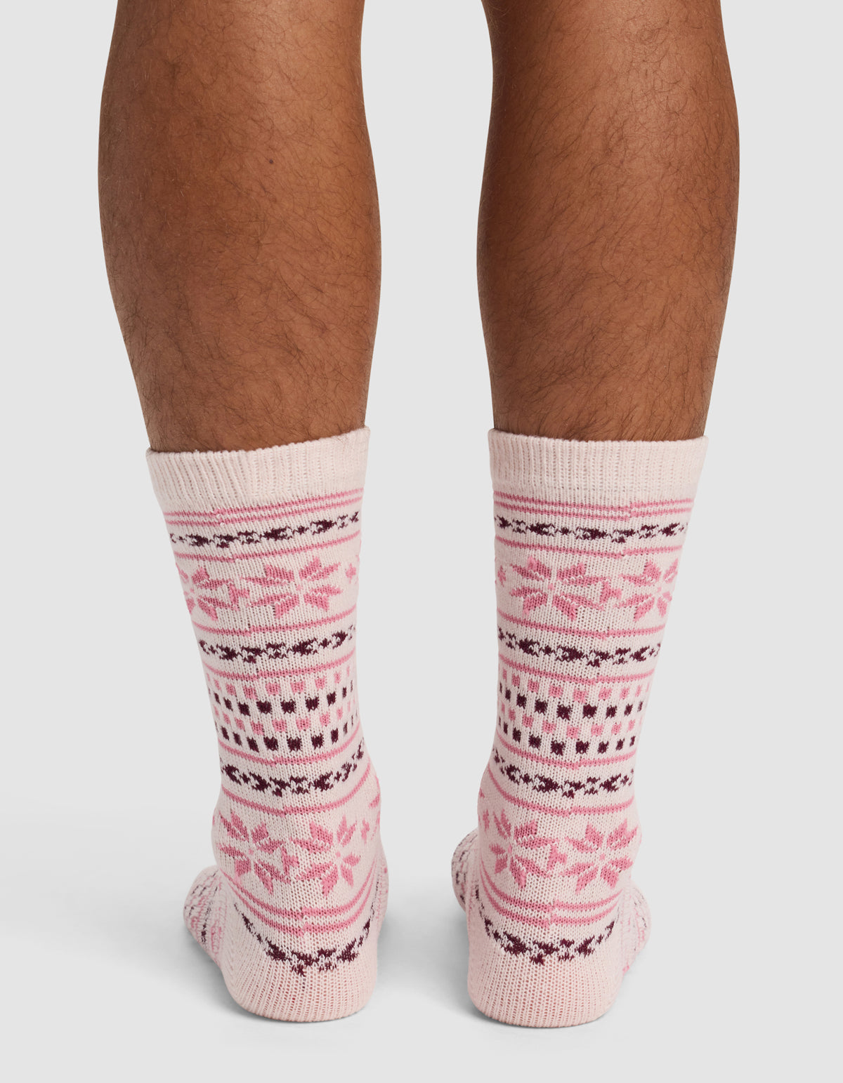 A person's lower legs and feet wear Cozy Earth's The Snowflake Plush Lounge Sock in light pink with dark pink and purple Nordic-inspired patterns, photographed from behind against a plain background.