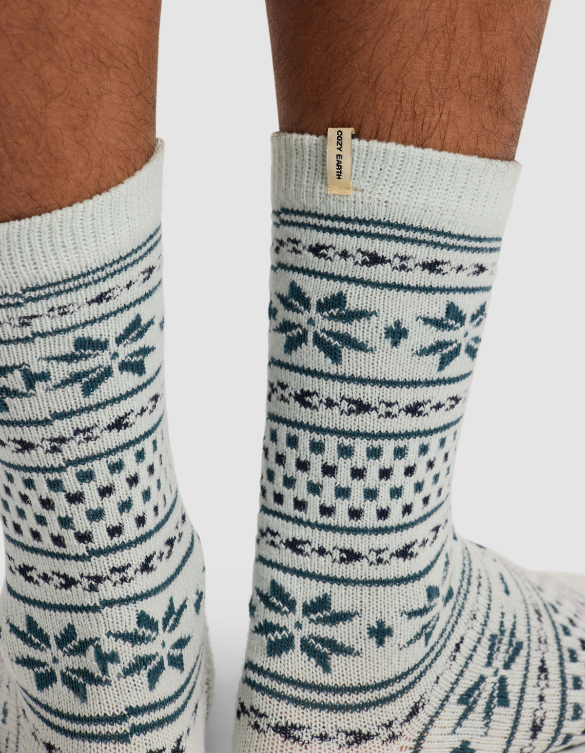 A close-up shows medium brown feet wearing Cozy Earth's The Snowflake Plush Lounge Sock—light-colored with blue Nordic-style patterns and a small beige tag.
