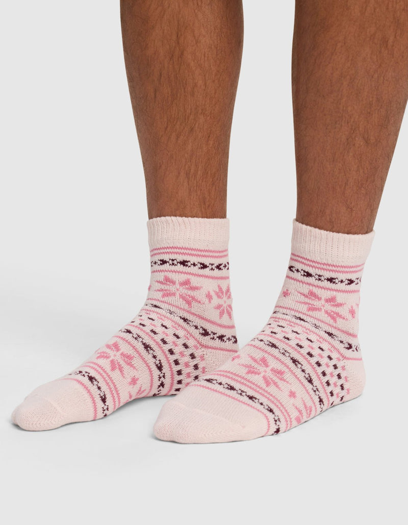 A person wears The Snowflake Plush Modern Crew Sock by Cozy Earth—light pink and white socks with dark pink and black snowflake and stripe patterns—standing on a white surface with only the lower legs and feet visible.