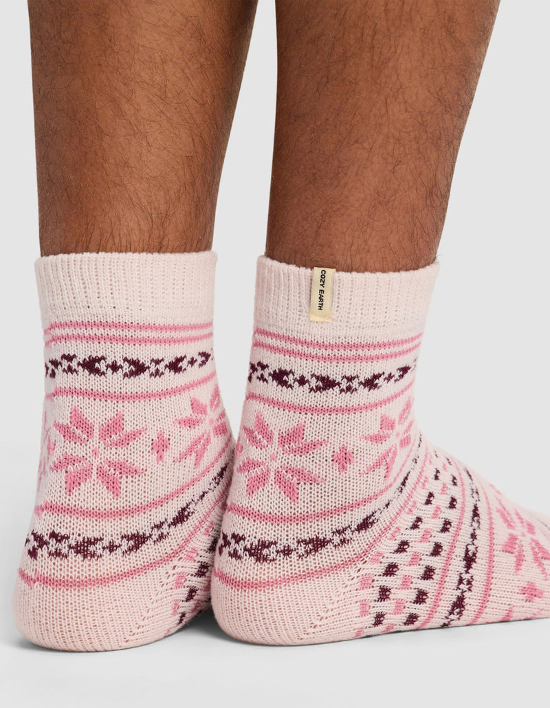 A close-up of feet wearing The Snowflake Plush Modern Crew Sock by Cozy Earth—pink, ankle-high socks with red and white snowflake patterns, a small beige tag near the cuff, set against a plain white background.
