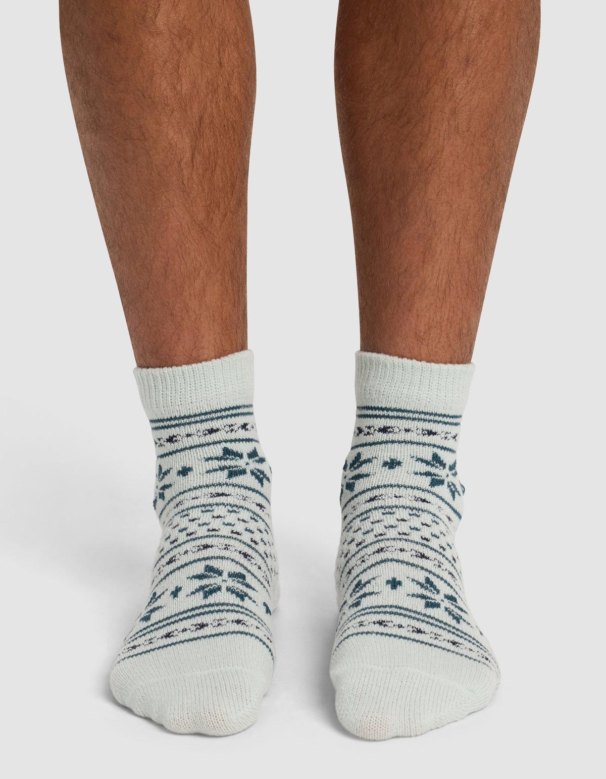 A person wearing Cozy Earth's The Snowflake Plush Modern Crew Sock—cream-colored with dark green and blue snowflake and stripe patterns—stands on a white surface.