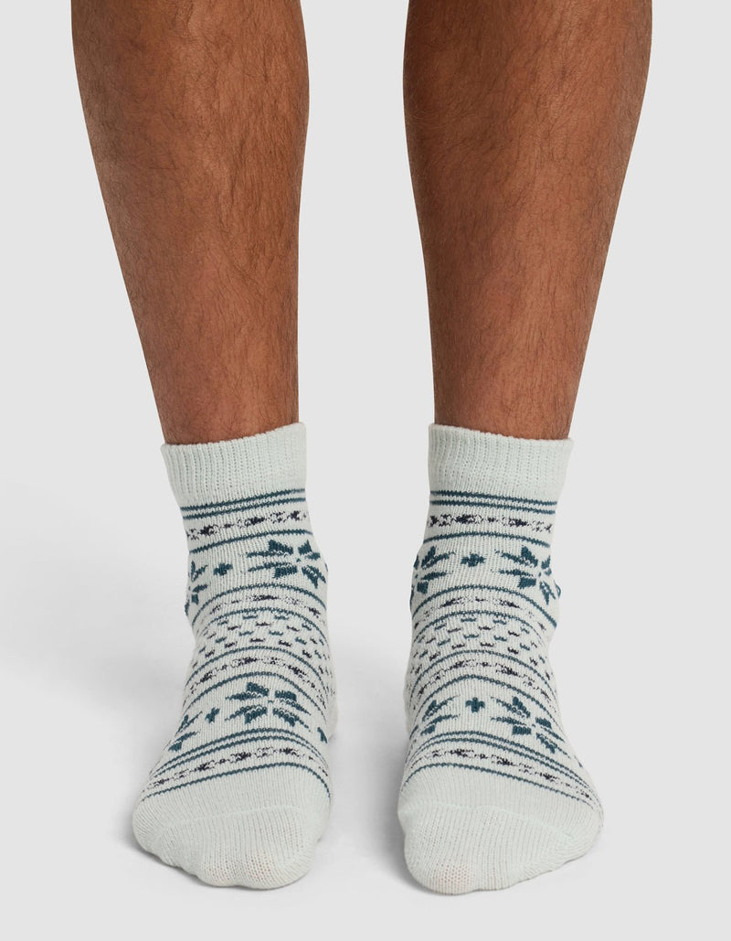 A person wearing Cozy Earth's The Snowflake Plush Modern Crew Sock—cream-colored with dark green and blue snowflake and stripe patterns—stands on a white surface.