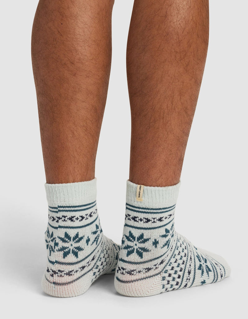 A person’s lower legs and feet are shown standing on a white surface, wearing Cozy Earth’s The Snowflake Plush Modern Crew Sock—light-colored socks with dark blue snowflakes and geometric patterns.