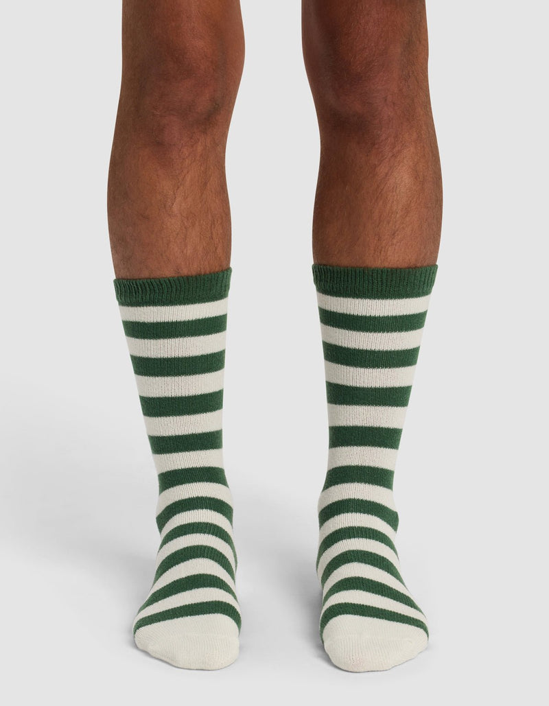 A person wearing The Striped Plush Lounge Sock by Cozy Earth, featuring knee-length horizontal green and white stripes, stands on a white background.