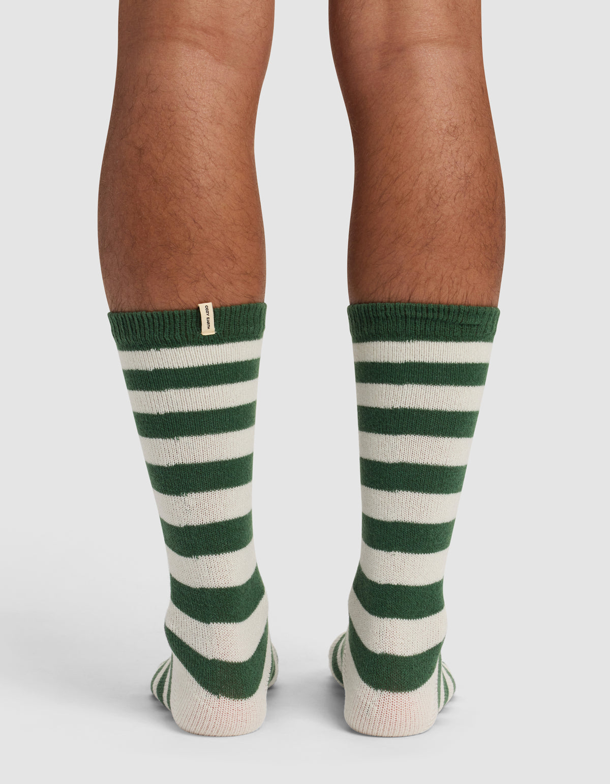 Back view of a person wearing Cozy Earth’s The Striped Plush Lounge Sock in green and white stripes, featuring a small beige tag near the top. The socks are shown on calves, standing on a light surface.|Color:Celadon Stripe
