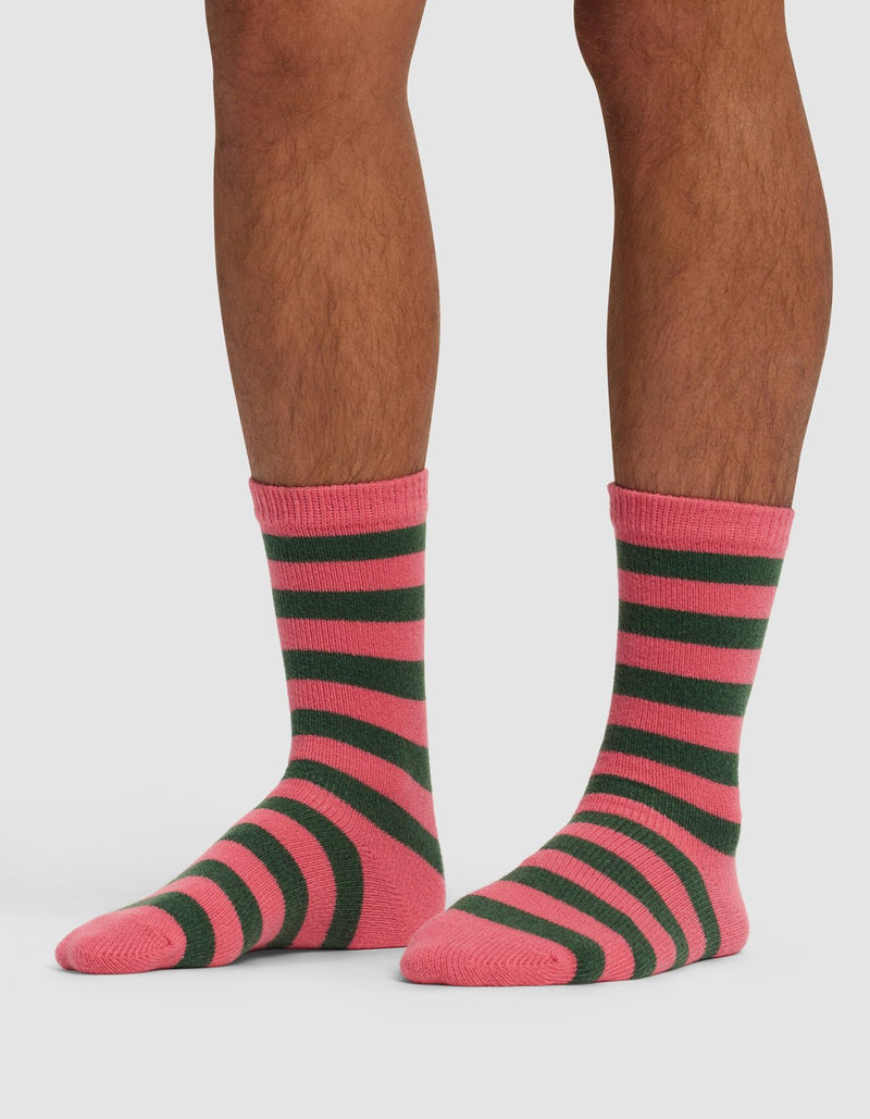 A person shows their lower legs and feet while wearing Cozy Earth's Striped Plush Lounge Sock in pink and dark green stripes, standing on a white background.