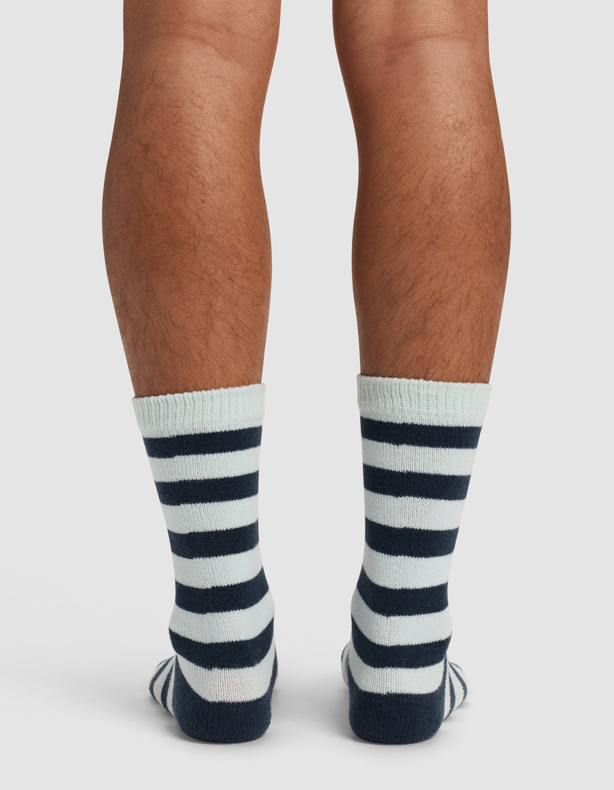 Back view of a person's lower legs wearing Cozy Earth's The Striped Plush Lounge Sock, featuring thick navy blue horizontal stripes on white, standing on a white background.