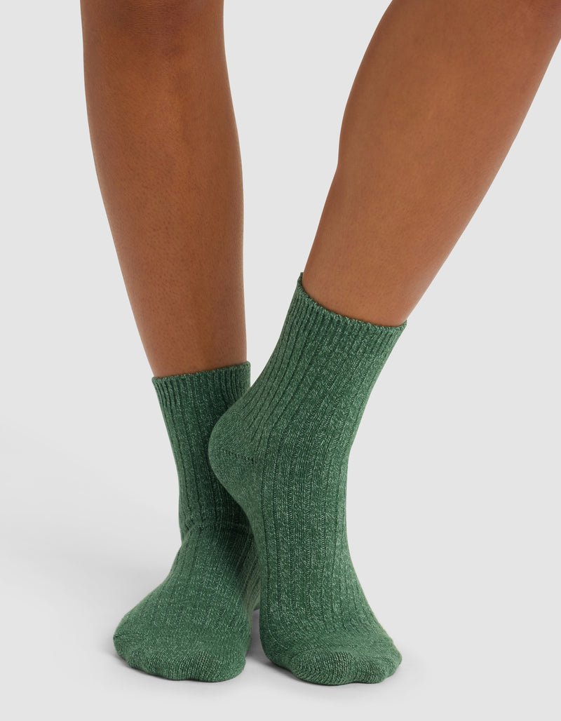 A person stands with feet crossed at the ankles, wearing Cozy Earth’s Plush Lounge Quarter Socks in green against a plain white background.
