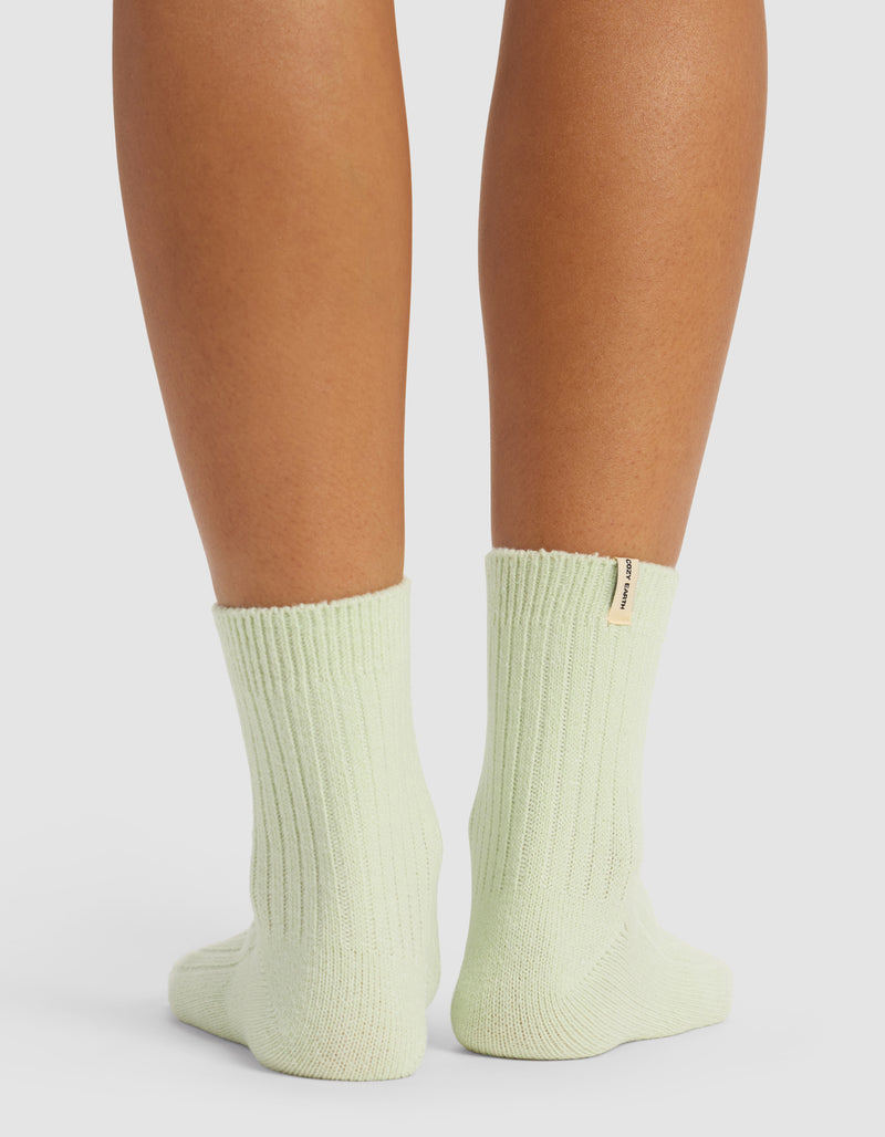 A close-up of a person’s lower legs in light green Plush Lounge Quarter Socks by Cozy Earth, featuring ribbed texture and a small tan tag on the right sock near the cuff, against a white background.