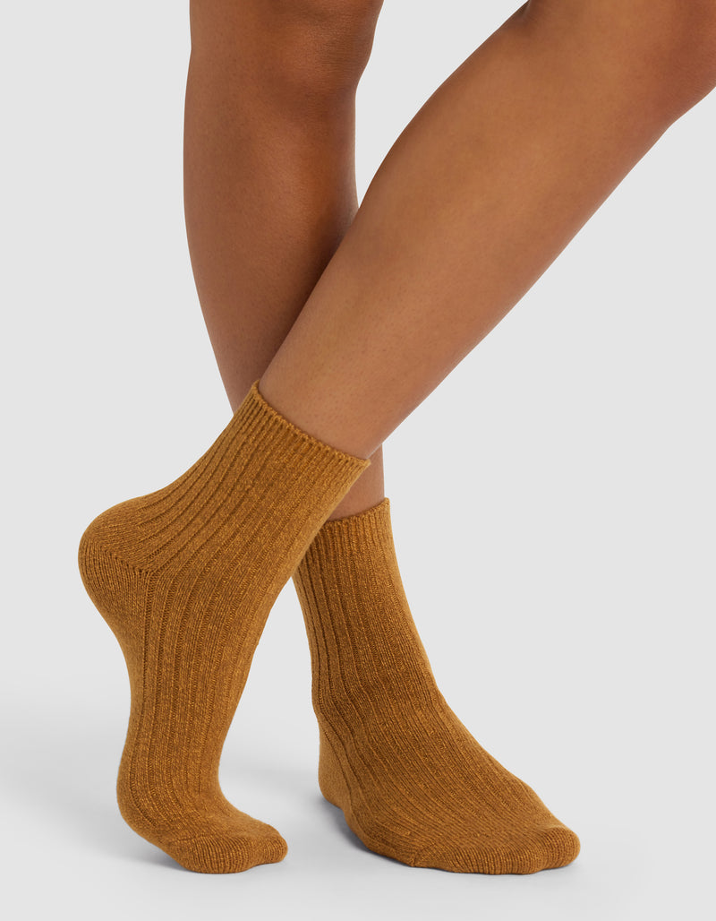 A person stands with legs crossed wearing Cozy Earth's Plush Lounge Quarter Socks 3-Pack in a thick, ribbed, mustard-colored style on a white background.
