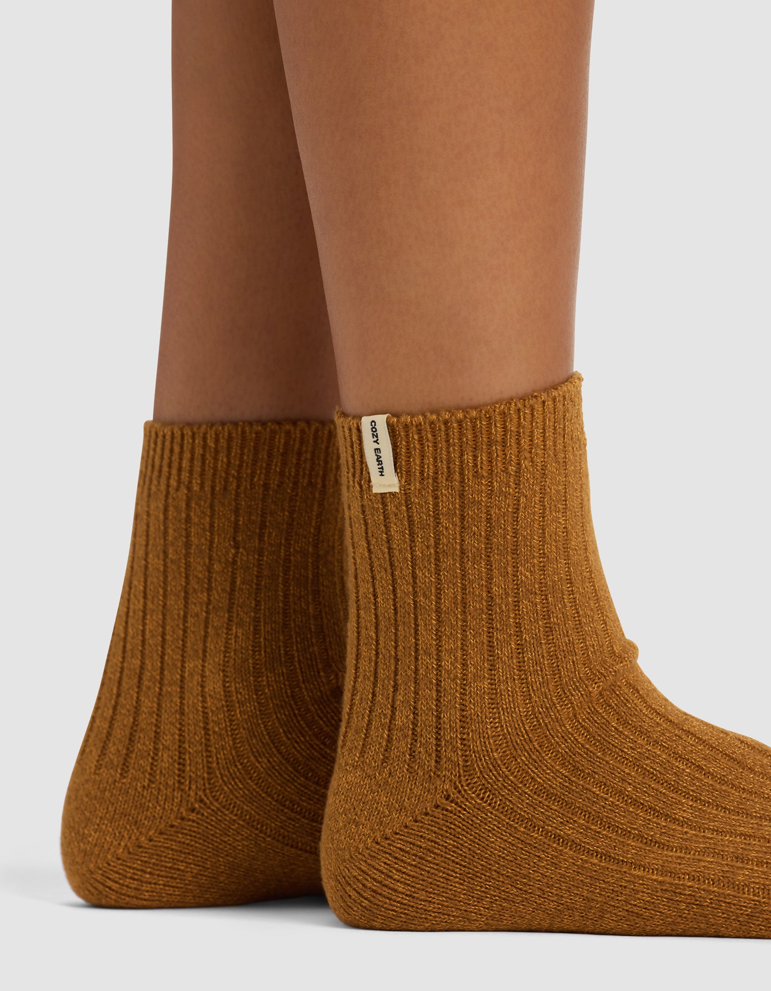 A close-up of two feet wearing Cozy Earth's Plush Modern Crew Socks in camel brown, featuring ribbed texture and a small beige side tag, shown against a plain background. Available as a 3-pack.