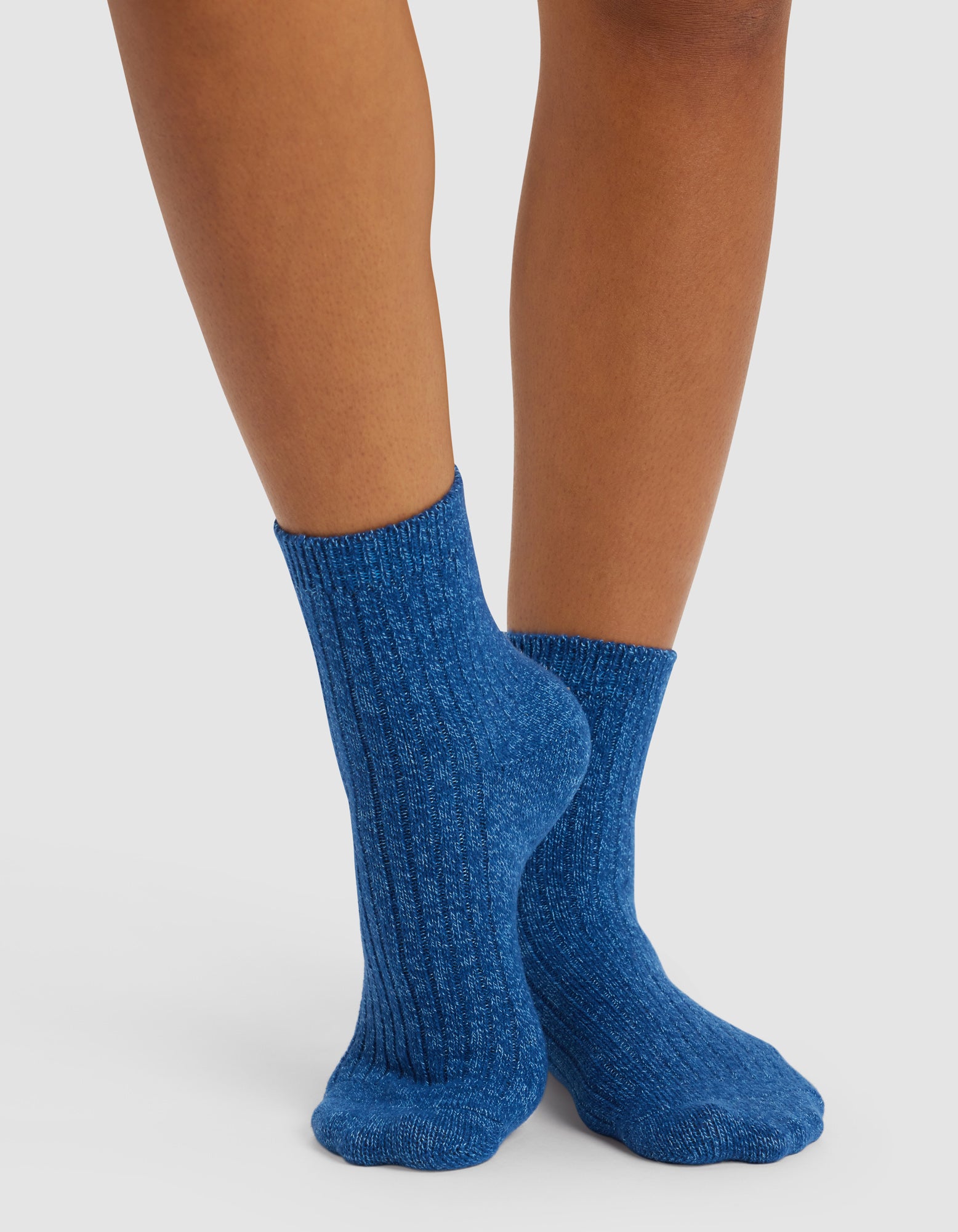 Someone wearing Cozy Earth’s Plush Lounge Quarter Sock 3-Pack in blue stands on a light surface with their feet crossed, showcasing the socks’ textured ribbed design.