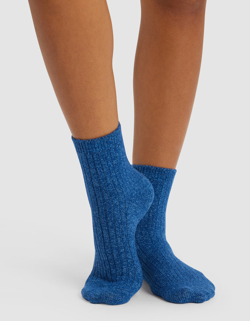 Someone wearing Cozy Earth’s Plush Lounge Quarter Sock 3-Pack in blue stands on a light surface with their feet crossed, showcasing the socks’ textured ribbed design.