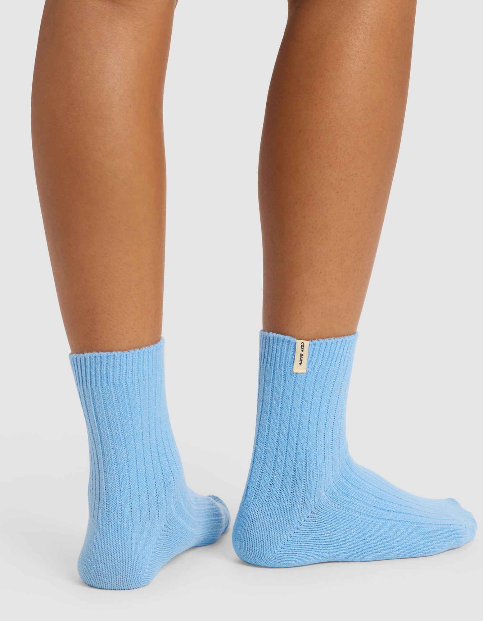 A person wears The Plush Modern Crew Sock - 3-Pack by Cozy Earth in light blue, featuring a small beige label on the back. Only the lower legs and feet are shown against a white background.