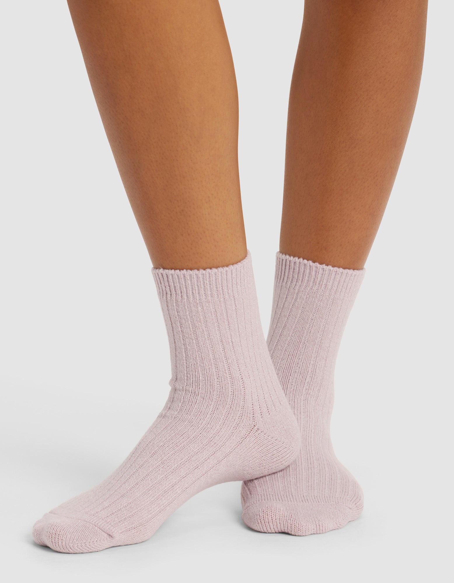 A person stands on a white surface wearing Cozy Earth’s The Plush Modern Crew Sock (3-Pack) in light pink; only their lower legs and feet are visible.