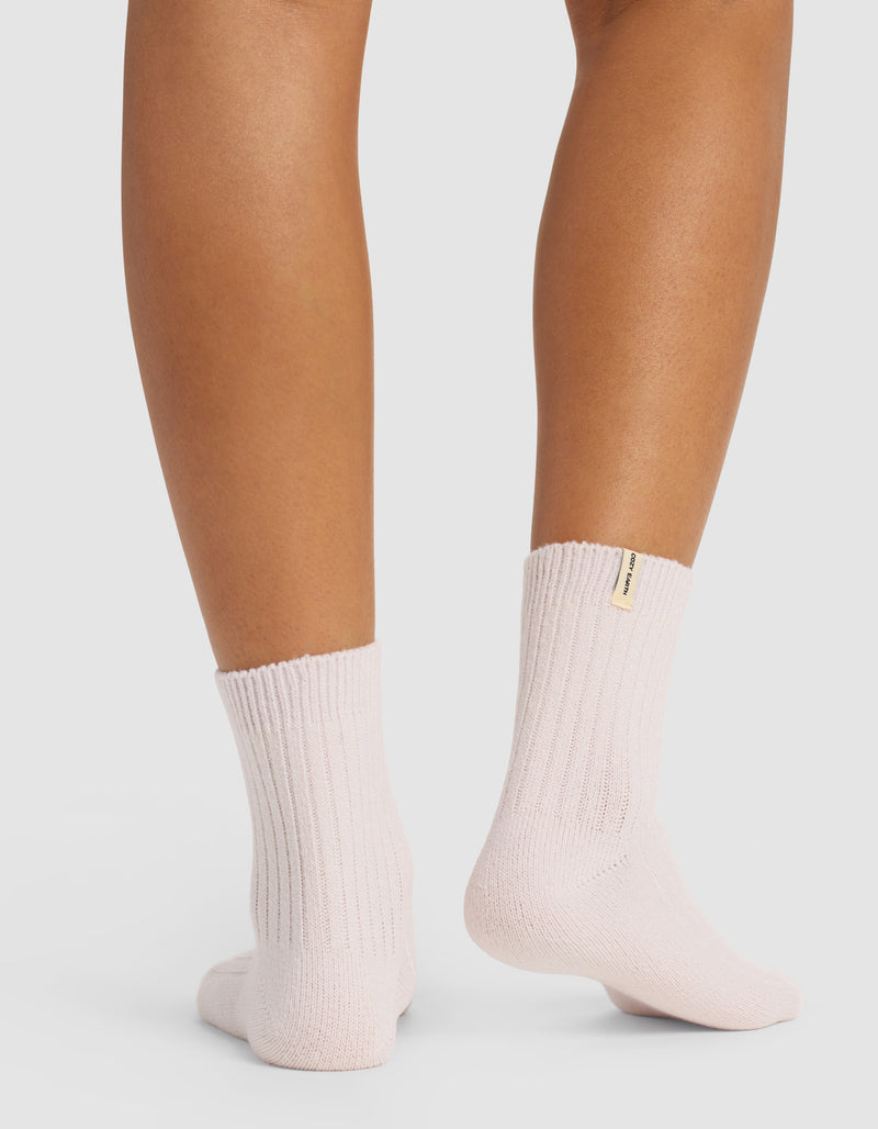 A person stands on a white surface, shown from the calves down, wearing light purple Plush Lounge Quarter Socks by Cozy Earth, featuring ribbed fabric and a small beige tag on one sock.