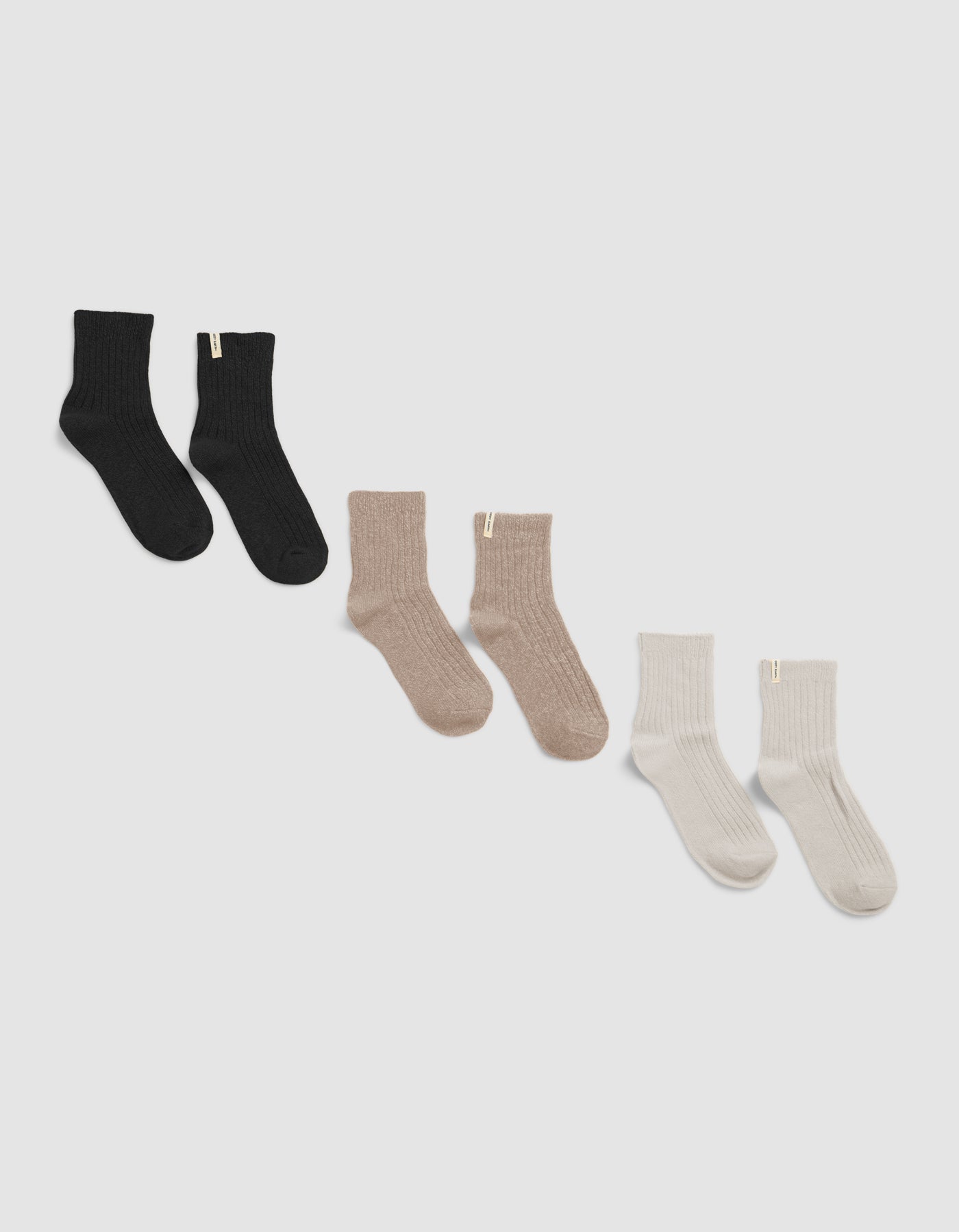 The Plush Modern Crew Sock 3-Pack by Cozy Earth is shown on a white background, featuring black, beige, and cream ribbed pairs, each with a small cuff tag.|Color:Black/Almond/Creme