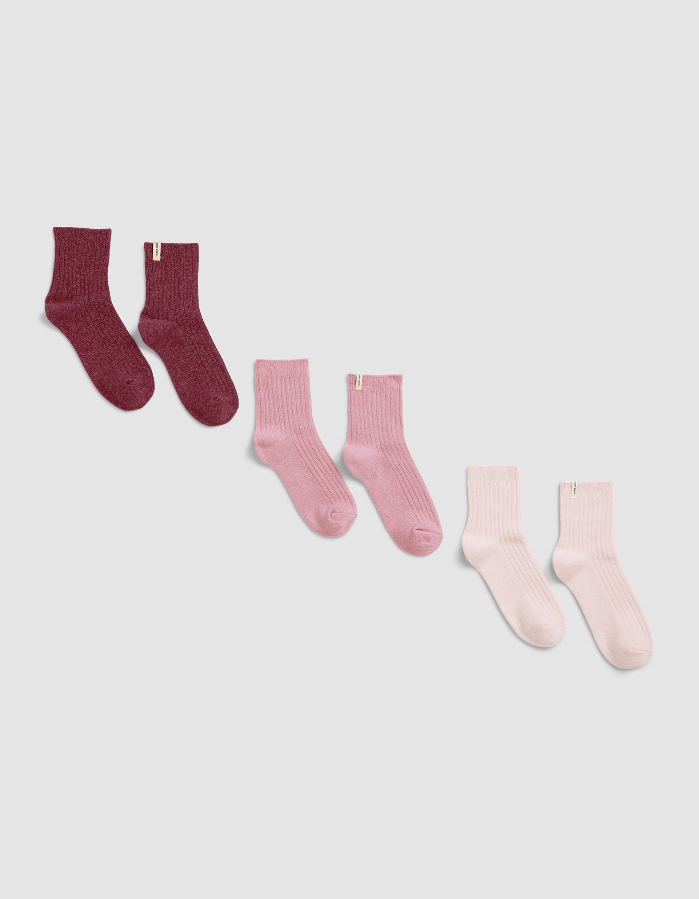 The Plush Modern Crew Sock 3-Pack by Cozy Earth, featuring ribbed socks in red, pink, and light pink, is displayed diagonally on a light gray background.