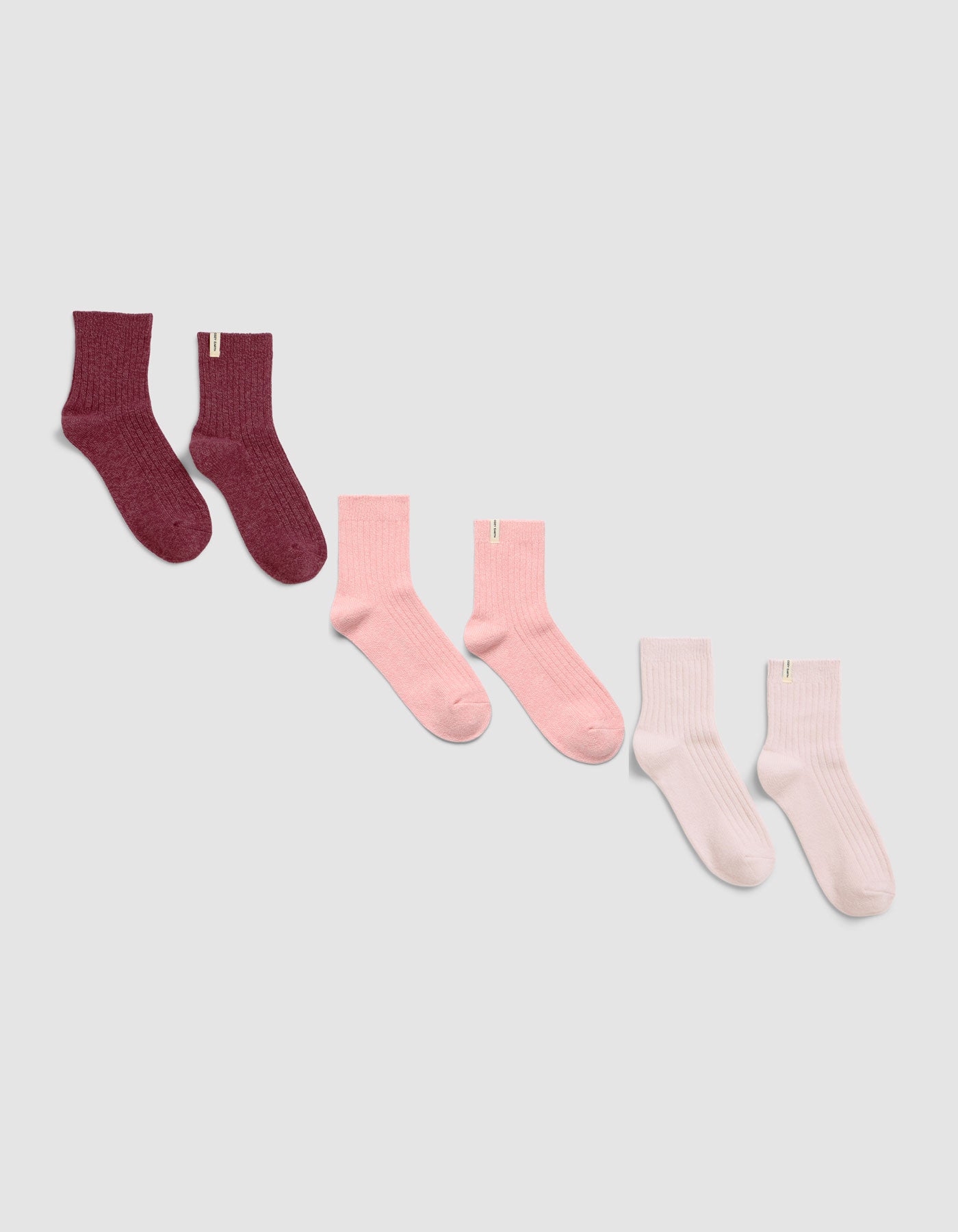 Three pairs of The Plush Modern Crew Sock 3-Pack by Cozy Earth, displayed diagonally on a light background in dark burgundy, pink, and light pink or white shades.|Color:Burgundy/Tulip/Blossom