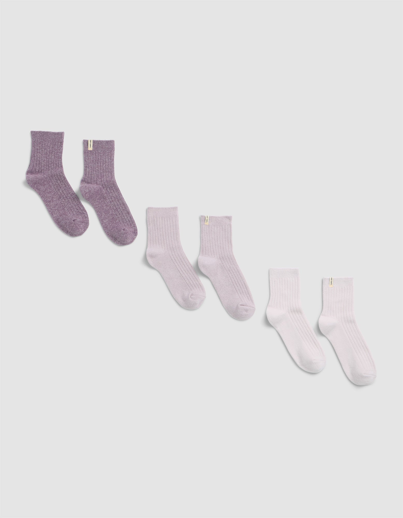 Three pairs of The Plush Modern Crew Sock by Cozy Earth, in purple, light lavender, and white, are displayed on a plain light gray background.
