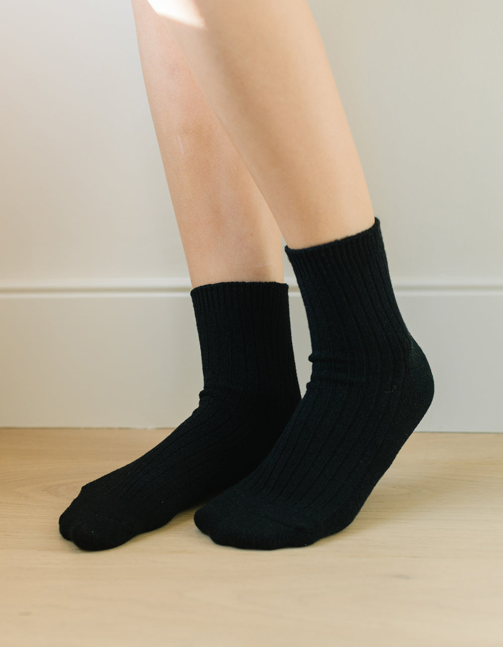 A person wearing The Plush Modern Crew Sock 3-Pack by Cozy Earth stands on a light wooden floor against a white wall.