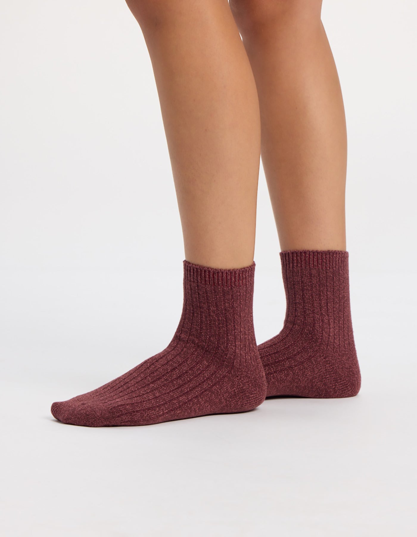 A person wearing maroon socks from the Cozy Earth Plush Modern Crew Sock 3-Pack stands on a white surface, with only their lower legs and feet visible.
